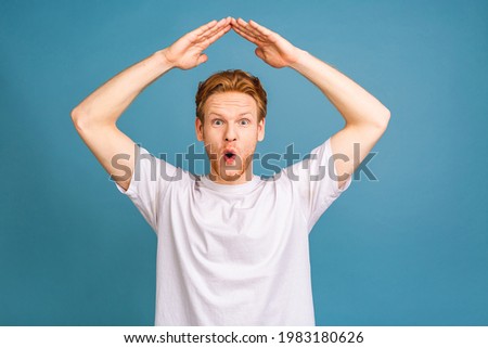 Happy satisfied handsome young man standing with roof gesture hands on head and looking with toothy smile. studio shot, isolated over blue background. Mortgage or finance concept