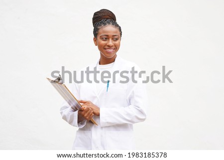 Young professional female black doctor with braided updo hair holds clipboard and wears a lab coat while smiling and looking to the right                                Royalty-Free Stock Photo #1983185378