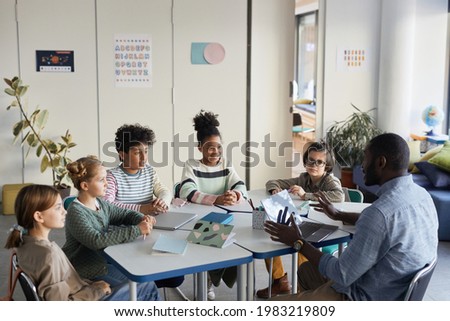 Diverse group of children sitting at table with male teacher in modern school classroom Royalty-Free Stock Photo #1983219809