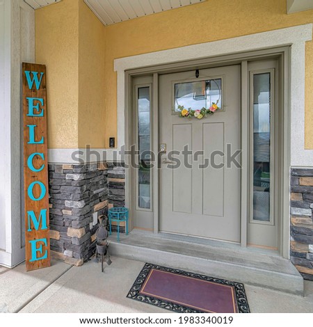 Square Gray front door with sidelights of a home with chairs at the concrete open porch. Floral door hanger and welcome sign board can also be seen at the entrance.