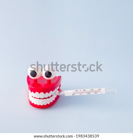 Plastic toy teeth with a thermometer in his mouth aganist pastel blue background. Abstract minimal healthy composition.