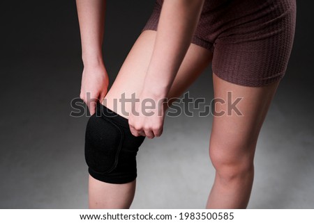 A girl pulls a knee pad on her leg before training in the gym after an injury. Isolated on gray background Royalty-Free Stock Photo #1983500585
