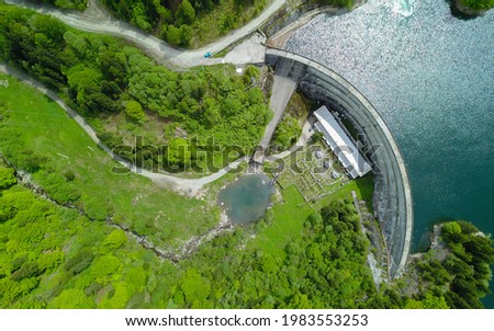 Aerial above panorama of Petrimanu Lake and a gravel road winding along its shores. The water is spilling over the dam, flowing through a concrete canal. Latorita Massif, Carpathia, Romania. Royalty-Free Stock Photo #1983553253