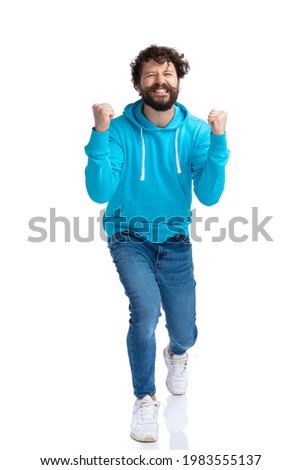 full body picture of excited bearded fashion model in blue hoodie holding arms up and celebrating victory, laughing and having fun on white background in studio