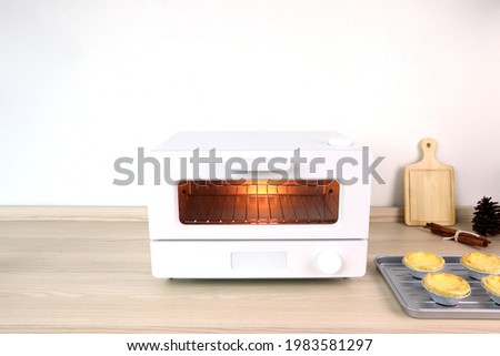 white modern toaster oven , countertop or convection oven is on the wooden table with homemade egg tarts , cinnamon stick and pinecone with white cement wall background in modern kitchen in morning