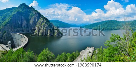 Lake Vidraru panorama. The arched, concrete dam, stops the flow of Capra stream, and creates a beautiful lake inside the mountains. Beech forest surround the blue waters. Fagaras, Carpathia, Romania. Royalty-Free Stock Photo #1983590174