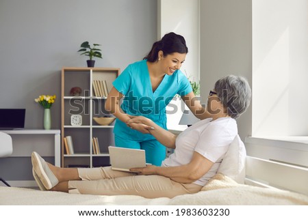 Happy elderly aged elegant woman patient lying on comfortable bed with book talking to female caregiver spending time together. Nursing home service and insurance healthcare for retired people Royalty-Free Stock Photo #1983603230