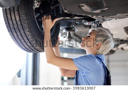 Caucasian female auto mechanic changing wheel tire in car in garage, side view. Beautiful young lady in overalls is concentrated on work, carefully adjusting repairing Royalty-Free Stock Photo #1983613007