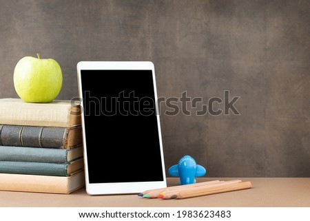 Back to school and education concept. The tablet with black screen, stack of books, apple, pens and plane near the blackboard and place for text. 