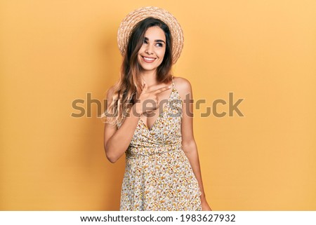 Young hispanic girl wearing summer hat cheerful with a smile of face pointing with hand and finger up to the side with happy and natural expression on face 