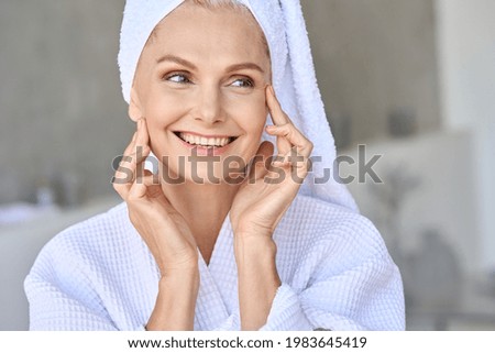 Portrait of happy attractive middle aged woman wearing bathrobe and white turban with bright complexion touching face looking away in bathroom. Advertising of skin care spa wellness concept. Royalty-Free Stock Photo #1983645419