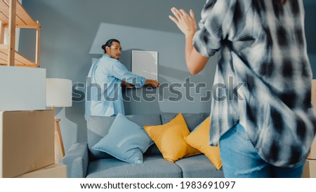 Happy asian young attractive couple man and woman help each other  hang picture frame on the wall decorate house and carton box package in living room. Young married asian decorating home concept. Royalty-Free Stock Photo #1983691097
