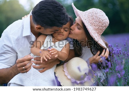 Young couple kissing little daughter sitting in lavender field and smiling. Charming baby girl wearing cute dress enjoying time with loving parents outdoors. Young family, nature concept.