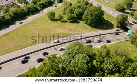 A high angle, aerial view over a parkway in New York, on a sunny day. There are green trees everywhere and the traffic is moving.
