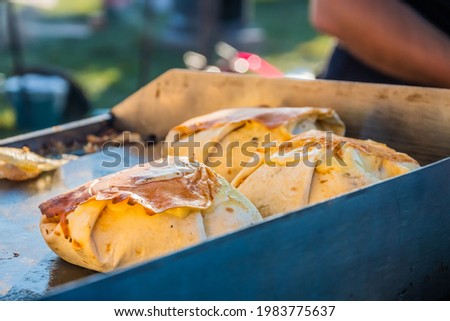Mexican food, a steak burger wrapped in a tortilla under melted cheese on the grill of an outdoor restaurant. Food festival in the city park. Street fast food.