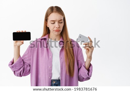 Doubtful young woman showing horizontal smartphone screen, looking skeptical at bank credit card, thinking, making choice, standing over white background