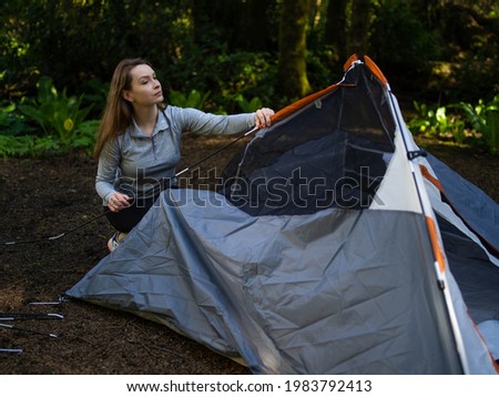 In the photo we see morning in the forest. Camping. Young woman folds up the tent. The woman looks up. In the background of the photo we see a dense forest.