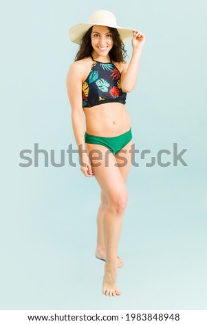 Hot attractive woman posing in a beautiful bikin against a blue background. Smiling young woman wearing a beach hat against a blue background