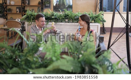 man with humor repeatedly photographs young smiling beautiful woman with coffee in her hands on cell phone camera in an outdoor cafe or restaurant.