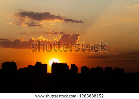 Sun sets just above high-rise buildings. Orange to white glow at its centre. Broad beams scatter and flare across sky. Clouds above orb. Buildings are in silhouette.