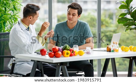 Male nutritionist showing and discussing vitamin pills with Asian man while sitting at table with healthy food while working in modern hospital Royalty-Free Stock Photo #1983915791