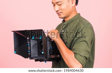 Portrait of Asian handsome young adult man confident professional videographer handheld holding digital video camera with matte box for cinema, studio shot isolated on pink background