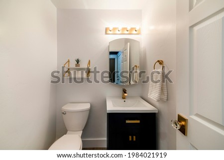 Modern powder room design with matching gold fixtures. Single vanity sink with dark wood cabinet and round mirror beside the toilet bowl under the layered shelves with gold brackets. Royalty-Free Stock Photo #1984021919