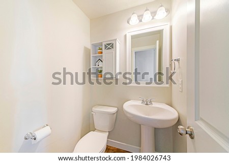 Interior of a white powder room with minimalist design. Toilet with ceramic bowl and sink with white wooden framed mirror beside the small white wooden cabinet and shelves with plants and house decor. Royalty-Free Stock Photo #1984026743