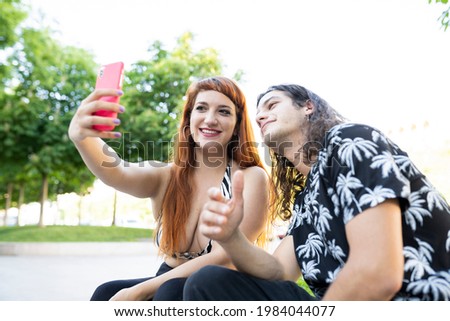 orange haired woman and long haired man sitting in a park taking a selfie