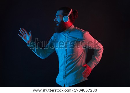 Photo of hipster man with beard wearing wireless headphones and running over dark background with neon light.