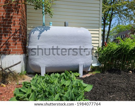 An oil tank hidden on the side of a suburban house in a close knit community. The tank is painted gray and nestled next to a brick chimney and flower beds filled with Hosta plants. Royalty-Free Stock Photo #1984077764