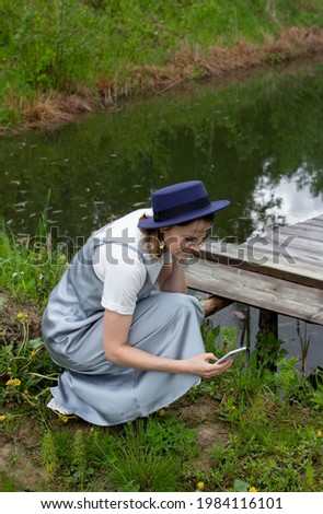 Young woman blogger sitting in the grass taking pictures with mobile phone, girl photographer with nature background, blonde in blue hat and long gray dress