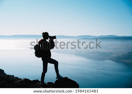 Side view of anonymous woman in casual clothes and with backpack standing on rocky edge and shooting picturesque background on camera