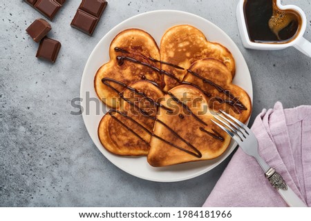 Pancakes in shape of breakfast hearts with chocolate sauce in gray ceramic plate, cup of coffee on gray concrete background. Table setting for Valentines Day breakfast. Top view copy space.