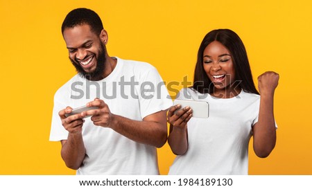 Online Battle Concept. Portrait of excited African american couple playing video game using smartphones, young woman making winner sign gesture, shaking fist, yellow orange studio background. Victory