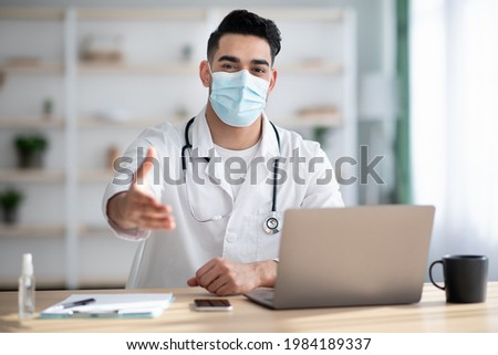 Young middle-eastern doctor in protective face mask sitting at workdesk with laptop at clinic, having appointment during coronavirus pandemic, greeting patient with outstretched hand