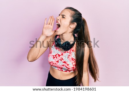Young brunette teenager wearing gym clothes and using headphones shouting and screaming loud to side with hand on mouth. communication concept. 