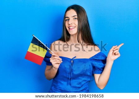 Young brunette teenager holding belgium flag smiling happy pointing with hand and finger to the side 