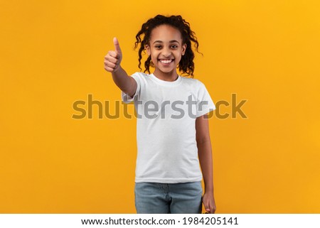 Best Choice, I Like It. Portrait of smiling black girl showing thumbs up sign gesture, approving or recommending something good. Teen posing isolated over orange yellow studio background
