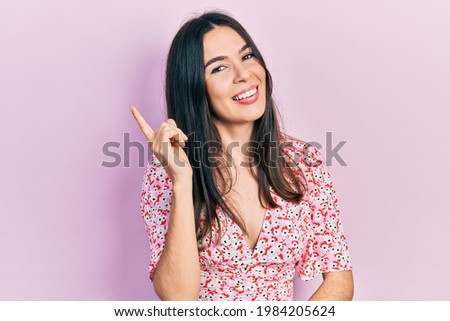 Young brunette woman wearing summer dress with a big smile on face, pointing with hand and finger to the side looking at the camera. 