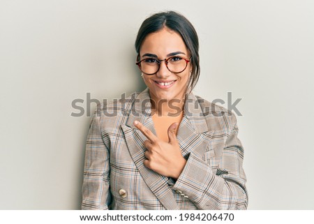 Young brunette woman wearing business jacket and glasses cheerful with a smile on face pointing with hand and finger up to the side with happy and natural expression 