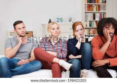 Bored group of multiracial friends relaxing at home sitting in a row on a comfortable sofa watching something off screen to the right with glum expressions Royalty-Free Stock Photo #198421205