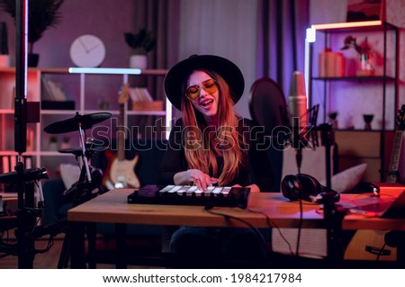 Female musician in trendy hat and glasses singing in microphone and using mixer console while recording music at home studio. Concept of people and talent. Royalty-Free Stock Photo #1984217984