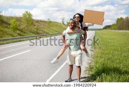 Millennial multiracial couple traveling by autostop, hitchhiking, looking for transport, trying to stop car, holding empty cardboard sign with mockup, ready strat new journey, copy space