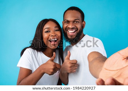 Super, We Like It. Portrait of smiling African American couple taking selfie and showing thumbs up sign gesture. Young guy and lady posing and looking at camera, isolated on blue studio background