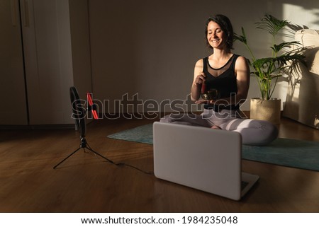 Happy young woman wearing sports clothes sitting at home and recording her yoga workout. She is holding a tibetan bowl.