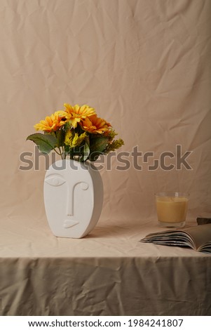 Still life photo of white oval ceramic vase shaped like human face. Modern creative flower vase with sunflowers is located on beige cloth near open book and aromatic candle.   Royalty-Free Stock Photo #1984241807
