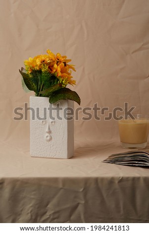 Still life photo of white rectangular ceramic vase shaped like human face. Modern creative flower vase with sunflowers is located on beige cloth near open book and aromatic candle.   Royalty-Free Stock Photo #1984241813