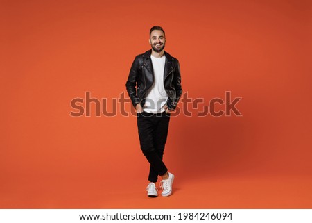 Full size length smiling cheerful young bearded man 20s wearing basic white t-shirt black leather jacket standing holding hands in pockets looking camera isolated on orange background studio portrait Royalty-Free Stock Photo #1984246094