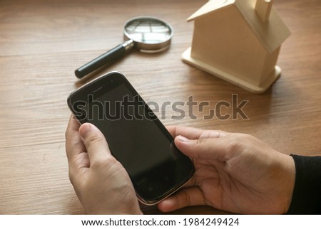 Hand holding a mobile phone searching for house with magnifying glass and house model at the background.